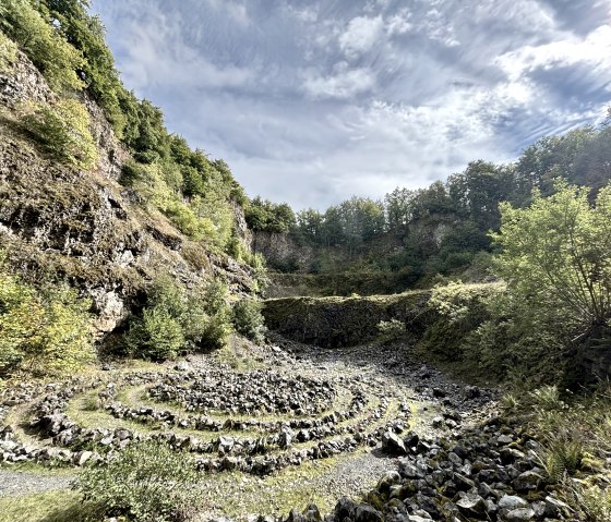 Steenspiraal in de Arensberg-vulkaan, omgeven door beboste rotswanden onder een blauwe lucht met wolken., © Touristik GmbH Gerolsteiner Land Steenspiraal in de Arensberg-vulkaan, omgeven door beboste rotswanden onder een blauwe lucht met wolken., © Touristik GmbH Gerolsteiner Land