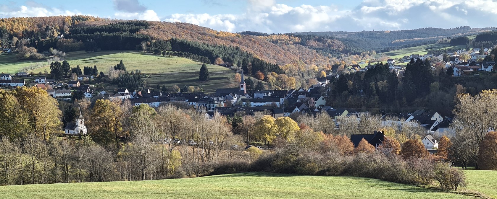 Panorama of Stadtkyll with green hills, autumnal trees and a church in the foreground. The sky is blue with a few clouds., © Touristik GmbH Gerolsteiner Land Panorama of Stadtkyll with green hills, autumnal trees and a church in the foreground. The sky is blue with a few clouds., © Touristik GmbH Gerolsteiner Land