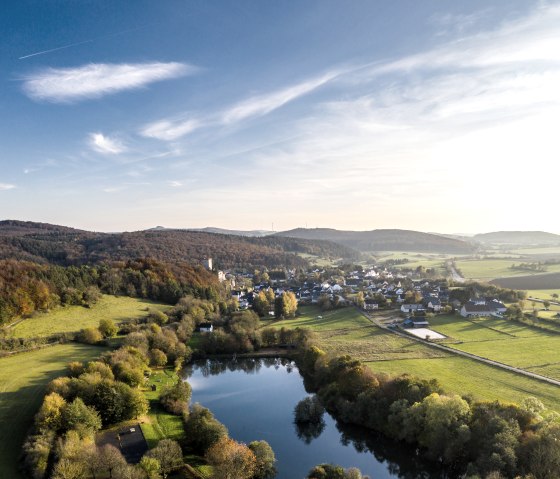 Aerial view of Kerpen in the Eifel: a lake surrounded by trees, a village in the background and rolling hills under a blue sky., © Eifel Tourismus GmbH, Dominik Ketz Aerial view of Kerpen in the Eifel: a lake surrounded by trees, a village in the background and rolling hills under a blue sky., © Eifel Tourismus GmbH, Dominik Ketz