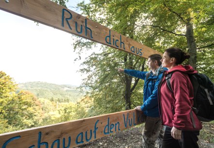 View of the Kalem volcano, Birresborn, © Eifel Tourismus GmbH, Dominik Ketz Two hikers stand at a viewpoint in a forest and point into the distance. They are standing by a wooden frame on which is written "Rest. View into the volcano".