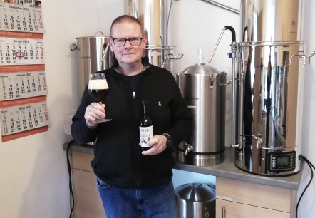 Herr Sibiak präsentiert sein Craftbeer "Dunkler Bock" vor seiner Brauanlage.
, © Touristik GmbH Gerolsteiner Land Ein Mann steht vor einer Brauanlage und hält eine Flasche Bier und ein gefülltes Glas hoch.