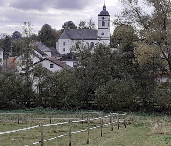 Kerk en huizen in een landelijke omgeving, omringd door bomen en weiden, onder een bewolkte hemel., © Touristik GmbH Gerolsteiner Land Kerk en huizen in een landelijke omgeving, omringd door bomen en weiden, onder een bewolkte hemel., © Touristik GmbH Gerolsteiner Land