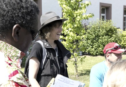 Führung "Verschwunden in der Krimistadt" Im Fokus eine dunkel gekleidete Frau mit Hut, die sehr lacht. Um Sie herum sind weitere Personen vor einem Gebäude.