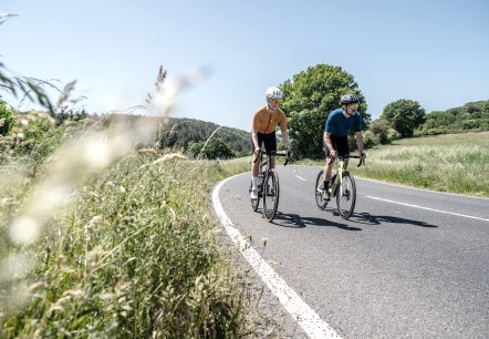 Twee fietsers rijden over een kronkelende landweg door een groen landschap in de Eifel. De lucht is helder en blauw., © Eifel Tourismus GmbH, Dennis Stratmann Twee fietsers rijden over een kronkelende landweg door een groen landschap in de Eifel. De lucht is helder en blauw., © Eifel Tourismus GmbH, Dennis Stratmann