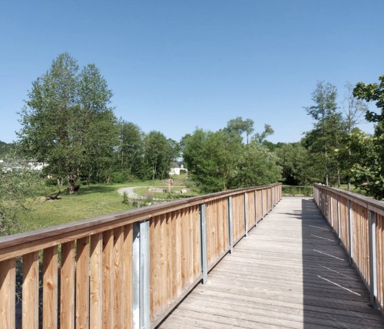Große Holzbrücke im Kurpark Stadtkyll, © Touristik GmbH Gerolsteiner Land Eine große Holzbrücke führt in ein Parkgelände mit zahlreichen Bäumen und Spazierwegen.