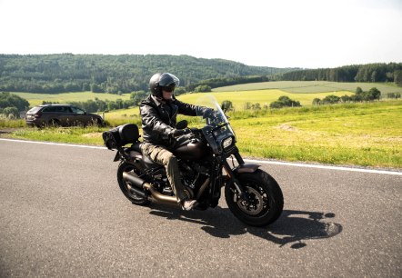 Motortocht door de Eifel
, © Thomas Hendele Een man in leren kleding en met een helm op zit op een motorfiets en rijdt op een weg naast een uitgestrekte weide. Aan de kant van de weg staat een auto geparkeerd.