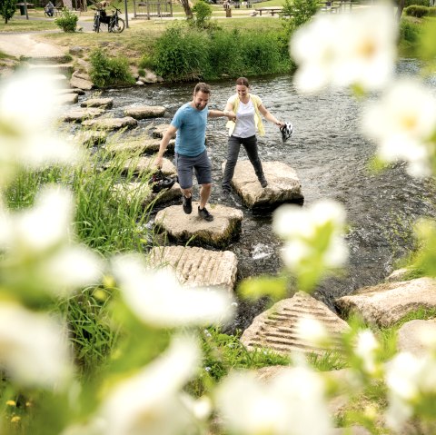 Two people cross the Kyll river in Gerolstein spa gardens using stepping stones.
, © Eifel Tourismus GmbH, Dominik Ketz A man and woman with helmets in their hands cross the Kyll river in Gerolstein spa gardens using stepping stones.