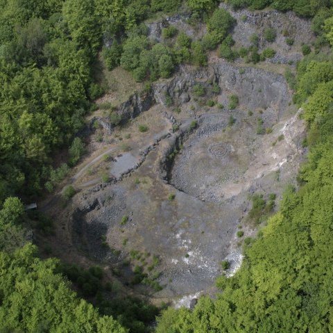 Aerial view of the forested crater of the Arensberg volcano with rocky areas and surrounding dense forest.
, © Thomas Regnery Aerial view of a forested crater with bare rocks and paths.