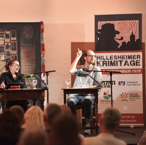 Die Krimistars Barbara Philipp, Steffen Will und Bernd Hölscher bei der Auftaktsveranstaltung der Hillesheimer Krimitage "Mord made in Eifel".
, © Helmut Gassen Zwei Männer und eine Frau sitzen nebeneinander an Tischen und lachen. Vor Ihnen dichtes Publikum und hinter Ihnen eine Indizienwand.