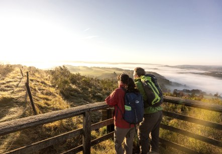Eifelsteig, Rother Kopf bij Müllenborn, © Eifel Tourismus GmbH, Dominik Ketz Twee wandelaars staan op een uitkijkplatform op de top van een berg en kijken uit over het herfstige en mistige landschap.