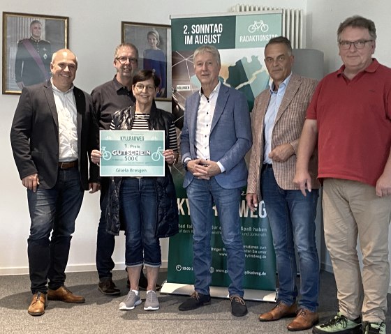 Preisverleihung Radaktionstag Grenzenlos Kyllradweg, v.l. Herr Hau (Westenergie), Herr Schmitz (Bürgermeister Stadtkyll), Frau Bresgen (Gewinnerin), Herr Wirtz (Bürgermeister Büllingen), Herr Westerburg (Bürgermeister Hellenthal), Herr Weicker (Bürgermeister Hallschlag), © Touristik GmbH Gerolsteiner Land Sechs Personen lächeln in die Kamera. Eine Frau hält einen großen Gutschein über 500 € in dne Händen.