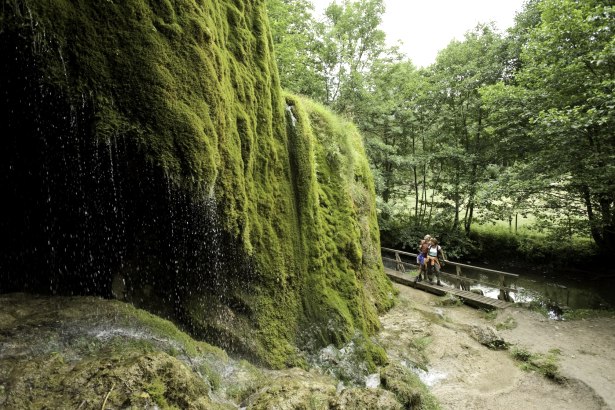 Der Nohner Wasserfall mit moosbewachsenen Felsen. Zwei Personen stehen auf einem Steg im Grünen. Ein idyllischer Zwischenstopp am Kalkeifel-Radweg., © Rheinland-Pfalz Tourismus GmbH/D. Ketz Der Nohner Wasserfall mit moosbewachsenen Felsen. Zwei Personen stehen auf einem Steg im Grünen. Ein idyllischer Zwischenstopp am Kalkeifel-Radweg., © Rheinland-Pfalz Tourismus GmbH/D. Ketz