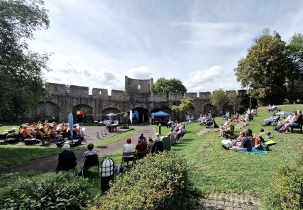 Event in the park area in front of the Hillesheim city wall
, © Touristik GmbH Gerolsteiner Land Numerous people are sitting on chairs and blankets on a lawn in the park, watching the small stage with the music association and readers in front of the Hillesheim city wall