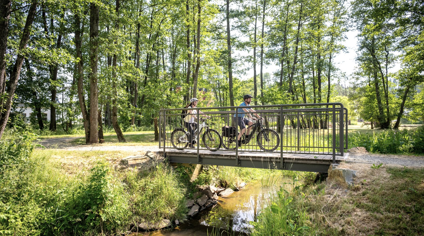 Two cyclists on the Kyll Cycle Route in Stadtkyll Spa Gardens.
, © Eifel Tourismus GmbH, Dominik Ketz A man and a woman ride e-bikes across a small bridge in the green Stadtkyll Spa Gardens.