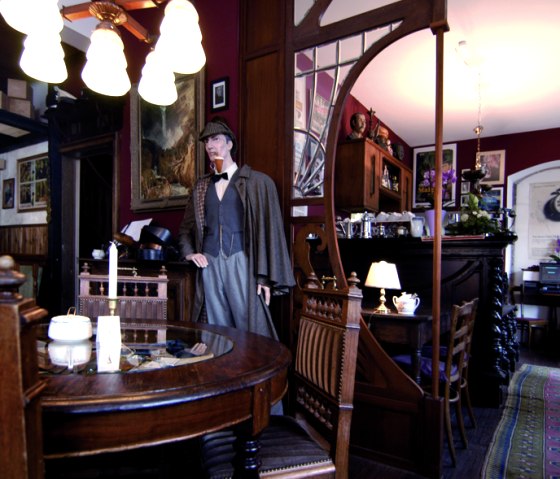A café with Victorian decor, a Sherlock Holmes figure, wooden furniture and cozy lighting. Pictures and a mirror hang on the wall., © Touristik GmbH Gerolsteiner Land A café with Victorian decor, a Sherlock Holmes figure, wooden furniture and cozy lighting. Pictures and a mirror hang on the wall., © Touristik GmbH Gerolsteiner Land