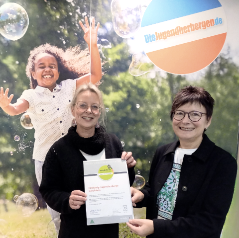 Das Foto zeigt zwei Frauen, die stolz ein Zertifikat halten, vor einem Hintergrund, der eine junge, fröhliche Person in Seifenblasen darstellt.Die Szene zeigt zwei Frauen, die sich für ein Foto aufgestellt haben, wobei eine das Zertifikat hält, das sie präsentieren. Der Hintergrund zeigt eine großformatige Darstellung eines Kindes, das inmitten von Seifenblasen fröhlich in die Kamera lächelt. Der Hintergrund ist ein grüner, naturähnlicher Hintergrund. Die Frauen stehen vor dem Hintergrund, wobei die Frau rechts das Zertifikat für die Kamera hält, während die Frau links ebenfalls beteiligt ist. Die Ausrichtung des Fotos ist gut und das Hauptaugenmerk liegt auf den Frauen und dem Zertifikat. Der Hintergrund dient als ergänzende Kulisse.Die beiden Hauptfiguren sind zwei Frauen mittleren Alters. Die Frau links hat blondes Haar, trägt eine Brille und einen schwarzen Pullover. Sie hält das Zertifikat mit einem Lächeln. Die Frau rechts hat kurz geschnittenes, braunes Haar, eine Brille und einen dunklen Blazer. Sie lächelt ebenfalls. Der Hintergrund zeigt ein glückliches Kind, das in Seifenblasen spielt., © Touristik GmbH Gerolsteiner Land Das Foto zeigt zwei Frauen, die stolz ein Zertifikat halten, vor einem Hintergrund, der eine junge, fröhliche Person in Seifenblasen darstellt.Die Szene zeigt zwei Frauen, die sich für ein Foto aufgestellt haben, wobei eine das Zertifikat hält, das sie präsentieren. Der Hintergrund zeigt eine großformatige Darstellung eines Kindes, das inmitten von Seifenblasen fröhlich in die Kamera lächelt. Der Hintergrund ist ein grüner, naturähnlicher Hintergrund. Die Frauen stehen vor dem Hintergrund, wobei die Frau rechts das Zertifikat für die Kamera hält, während die Frau links ebenfalls beteiligt ist. Die Ausrichtung des Fotos ist gut und das Hauptaugenmerk liegt auf den Frauen und dem Zertifikat. Der Hintergrund dient als ergänzende Kulisse.Die beiden Hauptfiguren sind zwei Frauen mittleren Alters. Die Frau links hat blondes Haar, trägt eine Brille und einen schwarzen Pullover. Sie hält das Zertifikat mit einem Lächeln. Die Frau rechts hat kurz geschnittenes, braunes Haar, eine Brille und einen dunklen Blazer. Sie lächelt ebenfalls. Der Hintergrund zeigt ein glückliches Kind, das in Seifenblasen spielt., © Touristik GmbH Gerolsteiner Land