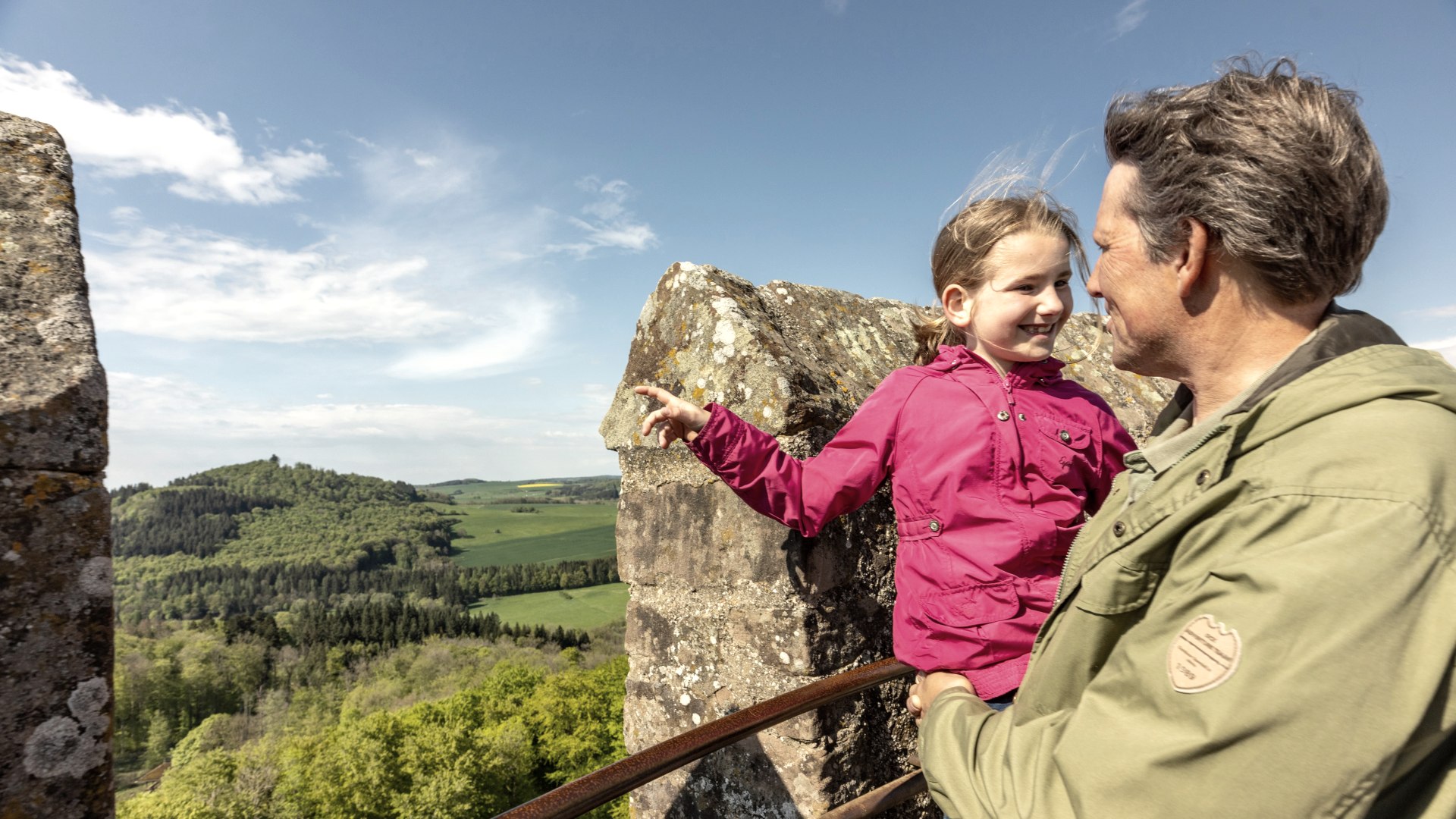 Adler & Wolfspark Kasselburg, Pelm, © Eifel Tourismus GmbH, Dominik Ketz Ein Vater trägt seine Tochter auf dem Arm auf einer Trumspitze einer Burg. Das Mädchen zeigt freudig in die Ferne.