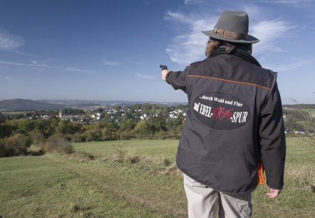 Willkomen im Krimiland!, © Molter Eine Hut tragende Person steht mit dem Rücken zum Betrachter und zeigt mit einer Pistole in die Ferne. Sie trägt eine Jacke mit der Aufschrift "...durch Wald und Flur auf Eifel-Krimi-Spur" auf dem Rücken. Vor der Person liegt eine große Wiese und dahinter in der Ferne ein Dorf mit vielen Häusern.