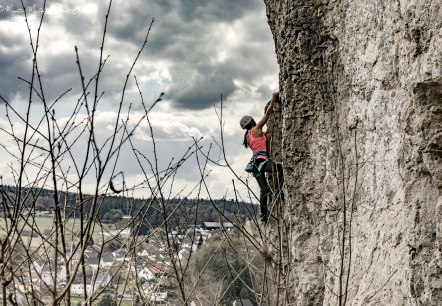 Een vrouw klimt op de steile rotswand van de Hustley. Op de achtergrond zijn de stad Gerolstein en een bewolkte hemel te zien.
, © Martin Müller Een vrouw, beveiligd met een helm en riemen, klimt op een steile rotswand. Op de achtergrond zijn een stad en een bewolkte hemel te zien.