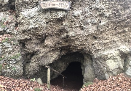 IJs- en molensteengrot "Martinshöhle" in Hohenfels-Essingen, © Touristik GmbH Gerolsteiner Land, Leonie Post Massieve rotswand met binnenin een kleine grotingang. Boven de grot hangt een bord met de naam "Martins-Höhle".