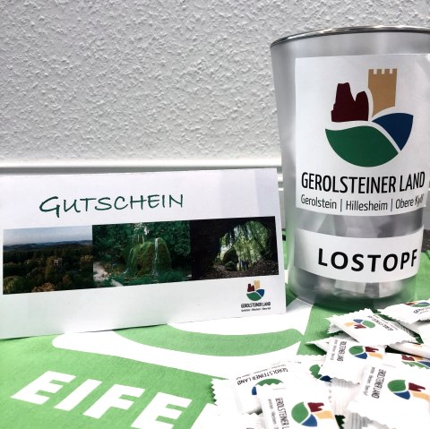 Gewinnspiel Facebook, © Touristik GmbH Gerolsteiner Land Ein Lostopf mit dem Logo der Ferienregion steht auf einem Tisch, daneben der zu gewinnende Reisegutschein und davor einige Traubenzucker.