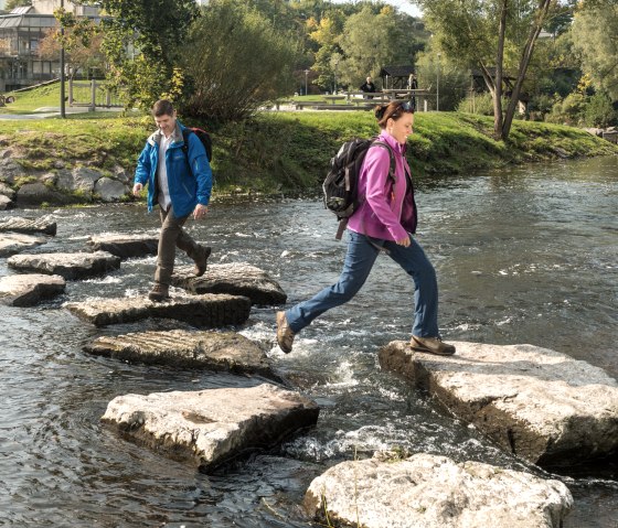 Wanderer überqueren die Kyll im Kurpark Gerolstein über Trittsteine.
, © Eifel Tourismus GmbH, Dominik Ketz Mann und Frau in Wanderkleidung springen über den Fluss Kyll im Kurpark Gerolstein über Trittsteine. Hinter Ihnen ist der Park mit Wasserspielplatz.