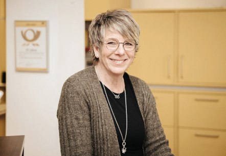 Jutta Perings, medewerkster van het postkantoor in Hillesheim, stelt zich voor.
, © Laura Kirwel Een vrouw staat achter de balie van een postkantoor. Achter haar staan kasten en het Posthorn-logo.