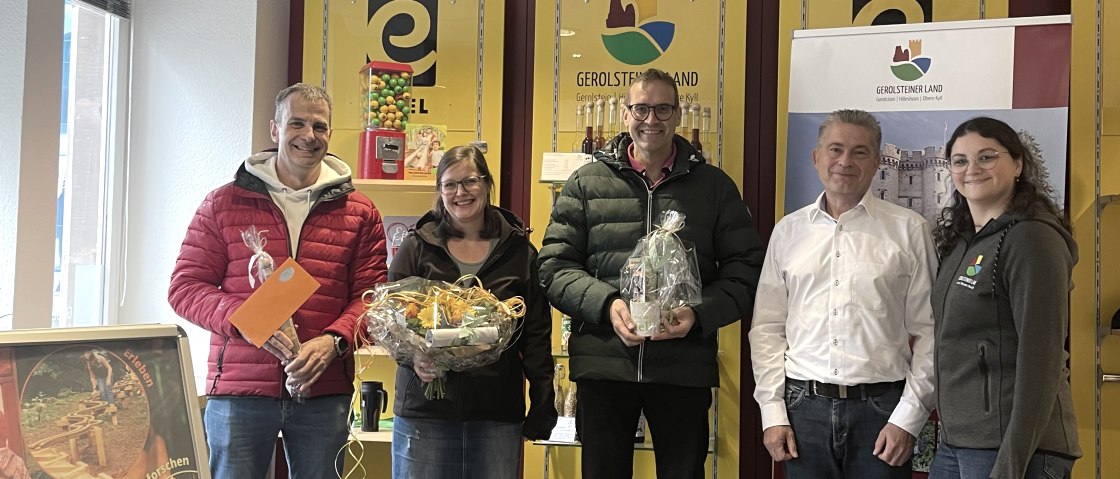 Frank Reuter, Geschäftsführer der Touristik GmbH Gerolsteiner Land, Mitarbeiterin Elvira Weimer und die 3 Gewinner des Gewinnspiels zum Eröffnungstag der Waldkugelbahn Gerolstein., © Touristik GmbH Gerolsteiner Land 5 Personen stehen nebeneinander vor einem Souvenierregal in der Tourist-Information Gerolstein und lachen in die Kamera. 3 der Personen halten stolz Preise in den Händen.