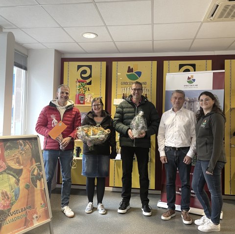 Frank Reuter, Geschäftsführer der Touristik GmbH Gerolsteiner Land, Mitarbeiterin Elvira Weimer und die 3 Gewinner des Gewinnspiels zum Eröffnungstag der Waldkugelbahn Gerolstein., © Touristik GmbH Gerolsteiner Land 5 Personen stehen nebeneinander vor einem Souvenierregal in der Tourist-Information Gerolstein und lachen in die Kamera. 3 der Personen halten stolz Preise in den Händen.