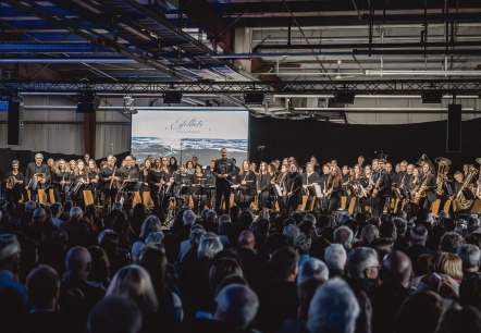 Konzert Eifelliebe
, © Eifelliebe Dunkle Halle, in der sich ein großes Orchester vor seinem Publikum erhebt.