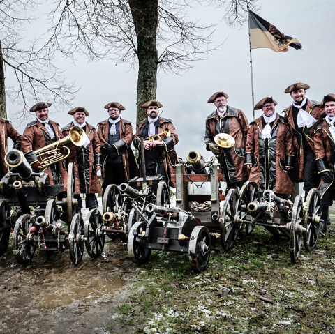 Auf einem Foto posiert eine Gruppe von Männern in historischer Kleidung, umgeben von kleinen Kanonen und Musikinstrumenten, vor dem Hintergrund einer trüben, winterlichen Landschaft.Die Szene zeigt eine Gruppe von neun Männern, die sich auf einer leicht erhöhten Grasfläche aufstellen, mit mehreren kleinen Kanonen vor sich und Musikinstrumenten wie Trompeten und einer Posaune. Sie scheinen entweder für ein Gruppenfoto zu posieren oder sich auf eine Vorführung vorzubereiten. Die Kanonen sind in einer Reihe angeordnet, mit einem dekorativen metallenen Hahn auf der linken Seite. Im Hintergrund ist eine bewölkte Landschaft mit Bäumen und einem Hügel mit einer Stadtansicht zu sehen, sowie eine Fahne mit einem Wappen. Die Komposition ist symmetrisch, wobei die Männer und Kanonen eine dominante horizontale Linie bilden, die durch die vertikalen Elemente der Bäume und die Fahne ausgeglichen wird.Die Hauptdarsteller sind neun Männer unterschiedlichen Alters, die traditionelle Kleidung aus dem 18. Jahrhundert tragen. Sie alle sind in braunen Röcken mit schwarzen Jacken, weißen Hemden und Halsbinden sowie Dreispitzenhüten gekleidet. Einige halten Musikinstrumente wie Trompeten und eine Posaune, während andere die Hände an ihren Hüften oder auf einem Gehstock ruhen lassen. Ihre Gesichtsausdrücke variieren, einige lächeln, während andere ernster blicken, was der Szene eine gewisse Lebendigkeit verleiht. Die Kanonen, die sie umgeben, sind detailreich gestaltet und aufwendig verziert, was auf ihre historische Bedeutung hinweist.Die fotografische Ausführung wirkt professionell, mit einem klaren Fokus auf die Hauptdarsteller und die Kanonen. Die Farbgebung ist natürlich und die Details sind gut erkennbar, was die Textur der Kleidung, der Kanonen und der Umgebung hervorhebt. Das Licht scheint von vorne zu kommen, was die Gesichter der Männer und die Details der Kanonen gut ausleuchtet. Der Stil der Fotografie ist dokumentarisch, was darauf hindeutet, dass das Foto wahrscheinlich bei einer Veranstaltung oder einem historischen Reenactment aufgenommen wurde.Die Szene spielt sich an einem bewölkten Tag ab, was eine eher gedämpfte und kühle Atmosphäre erzeugt. Der Hintergrund ist verschwommen, was die Hauptdarsteller hervorhebt, während die Bäume und die Stadtansicht im Hintergrund einen Kontext für die Szene schaffen. Der Boden ist mit nassem Gras bedeckt, was auf die Jahreszeit hindeutet und die raue Natur der Umgebung verstärkt. Die leicht getrübte Beleuchtung und der Hintergrund schaffen eine gedämpfte, realistische Atmosphäre, die gut zur historischen Thematik passt., © Klaus Wollwert Auf einem Foto posiert eine Gruppe von Männern in historischer Kleidung, umgeben von kleinen Kanonen und Musikinstrumenten, vor dem Hintergrund einer trüben, winterlichen Landschaft.Die Szene zeigt eine Gruppe von neun Männern, die sich auf einer leicht erhöhten Grasfläche aufstellen, mit mehreren kleinen Kanonen vor sich und Musikinstrumenten wie Trompeten und einer Posaune. Sie scheinen entweder für ein Gruppenfoto zu posieren oder sich auf eine Vorführung vorzubereiten. Die Kanonen sind in einer Reihe angeordnet, mit einem dekorativen metallenen Hahn auf der linken Seite. Im Hintergrund ist eine bewölkte Landschaft mit Bäumen und einem Hügel mit einer Stadtansicht zu sehen, sowie eine Fahne mit einem Wappen. Die Komposition ist symmetrisch, wobei die Männer und Kanonen eine dominante horizontale Linie bilden, die durch die vertikalen Elemente der Bäume und die Fahne ausgeglichen wird.Die Hauptdarsteller sind neun Männer unterschiedlichen Alters, die traditionelle Kleidung aus dem 18. Jahrhundert tragen. Sie alle sind in braunen Röcken mit schwarzen Jacken, weißen Hemden und Halsbinden sowie Dreispitzenhüten gekleidet. Einige halten Musikinstrumente wie Trompeten und eine Posaune, während andere die Hände an ihren Hüften oder auf einem Gehstock ruhen lassen. Ihre Gesichtsausdrücke variieren, einige lächeln, während andere ernster blicken, was der Szene eine gewisse Lebendigkeit verleiht. Die Kanonen, die sie umgeben, sind detailreich gestaltet und aufwendig verziert, was auf ihre historische Bedeutung hinweist.Die fotografische Ausführung wirkt professionell, mit einem klaren Fokus auf die Hauptdarsteller und die Kanonen. Die Farbgebung ist natürlich und die Details sind gut erkennbar, was die Textur der Kleidung, der Kanonen und der Umgebung hervorhebt. Das Licht scheint von vorne zu kommen, was die Gesichter der Männer und die Details der Kanonen gut ausleuchtet. Der Stil der Fotografie ist dokumentarisch, was darauf hindeutet, dass das Foto wahrscheinlich bei einer Veranstaltung oder einem historischen Reenactment aufgenommen wurde.Die Szene spielt sich an einem bewölkten Tag ab, was eine eher gedämpfte und kühle Atmosphäre erzeugt. Der Hintergrund ist verschwommen, was die Hauptdarsteller hervorhebt, während die Bäume und die Stadtansicht im Hintergrund einen Kontext für die Szene schaffen. Der Boden ist mit nassem Gras bedeckt, was auf die Jahreszeit hindeutet und die raue Natur der Umgebung verstärkt. Die leicht getrübte Beleuchtung und der Hintergrund schaffen eine gedämpfte, realistische Atmosphäre, die gut zur historischen Thematik passt., © Klaus Wollwert