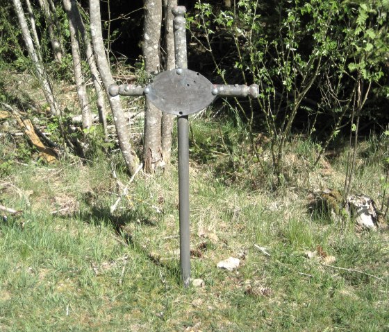 Een zwart metalen kruis met een ovale plaat staat in een weiland omringd door bomen en struiken., © Touristik GmbH Gerolsteiner Land Een zwart metalen kruis met een ovale plaat staat in een weiland omringd door bomen en struiken., © Touristik GmbH Gerolsteiner Land