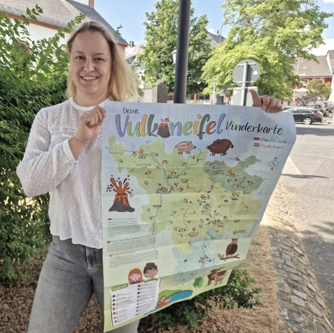 Mitarbeiterin Esther Erharter präsentiert die neue Vulkaneifel Kinderkarte.
, © Touristik GmbH Gerolsteiner Land Eine Frau steht auf einem Platz vor einer Hecke und hält eine große Karte mit Überschrift "Vulkaneifel Kinderkarte" hoch.