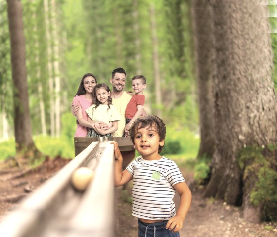 Familie an der Waldkugelbahn, © Kurt Daun Eltern mit zwei Kindern auf dem Arm stehen hinter einer Hoz-Kugelbahn und ein Kind steht daneben im Wald.
