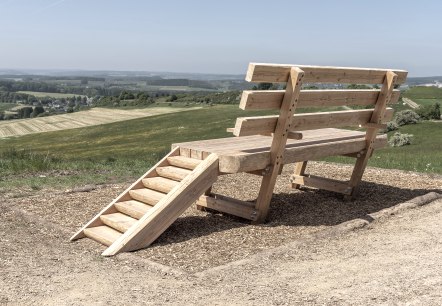 Een oversized houten rustbank staat op een heuvel voor een uitgestrekt landschap., © Jochen Hank Een oversized houten rustbank staat op een heuvel voor een uitgestrekt landschap., © Jochen Hank