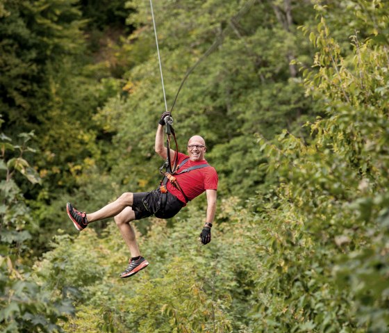 Ein Mann in rotem Shirt und schwarzer Shorts fährt lachend auf einer Zipline durch einen grünen Wald., © Eifel-Adventure, Andreas Goltz