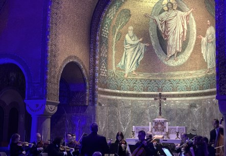Zwak verlicht interieur van de Erl&ouml;serkirche in Gerolstein met gouden moza&iuml;ekversieringen. In het altaargedeelte muzikanten en een groot publiek op de banken.