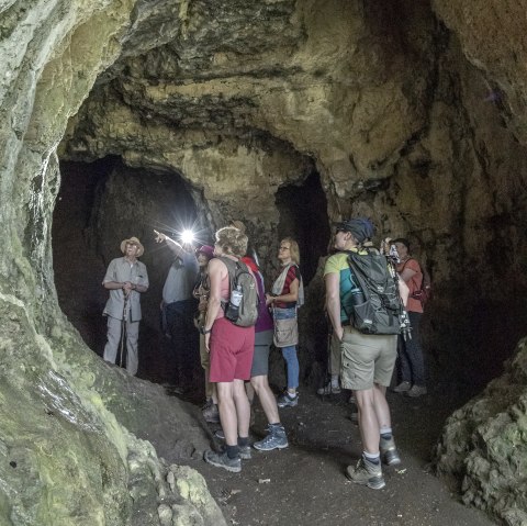 A hiking group of around 10 people stands in the interior of a large cave. The people are marvelling at the massive stone walls around them.