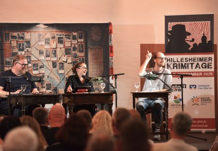 Zwei Männer und eine Frau sitzen nebeneinander an Tischen und lachen. Vor Ihnen dichtes Publikum und hinter Ihnen eine Indizienwand.
