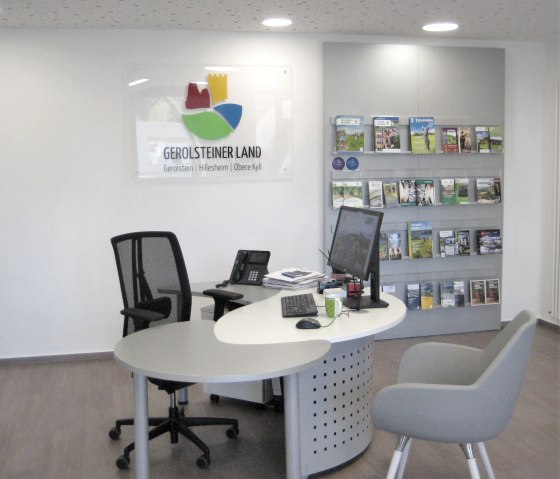 Innenraum der Tourist-Information Stadtkyll mit Arbeitsplatz inkl. Computer, Telefon, Tastatur etc., daneben eine Regalwand mit Pospekten.