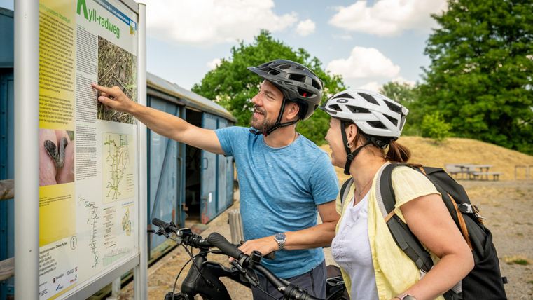 Twee fietsers met helmen kijken naar een informatiebord langs de Kyll-fietsroute. De man wijst naar het bord, terwijl de vrouw glimlachend toekijkt. Fietsen zijn op de voorgrond.