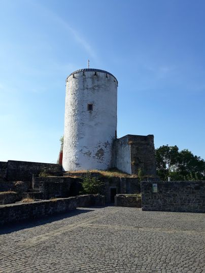 Ein alter runder Turm mit einer weißen Fassade steht auf einer befestigten Fläche. Im Hintergrund ist blauer Himmel zu sehen.