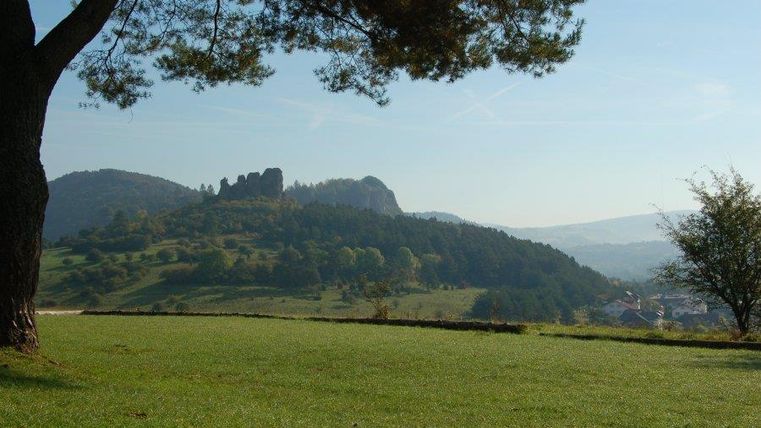 Eine malerische Landschaft mit sanften Hügeln und einem klaren Himmel. Im Hintergrund sind Ruinen sichtbar, umgeben von grünen Wiesen.