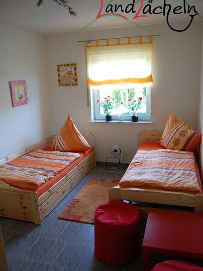 Ein gemütliches Zimmer mit zwei Einzelbetten und oranger Bettwäsche. Der Raum hat ein Fenster mit Blick auf die Pflanzen draußen und einen roten Hocker.