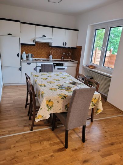 Une cuisine moderne avec une grande table et six chaises. La table est couverte d'une nappe fleurie et les meubles de cuisine sont principalement blancs.
