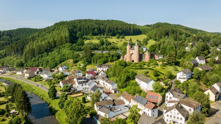 Luftaufnahme der Bertradaburg in Mürlenbach, umgeben von grünen Hügeln und einem kleinen Dorf. Ein Fluss fließt durch die Landschaft.