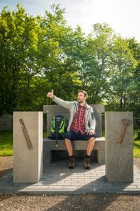 Ein mann in Wanderkleidung und mit Rucksack sitzt auf einem Steinthron auf einer Anhöhe vor Bäumen. Er schaut in eine Richtung, lacht und hebt den Daumen.