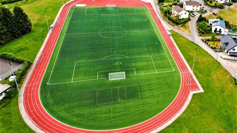 Ein Sportplatz mit Kunstrasen und einer Laufbahn. Umgeben von Wohnhäusern und Grünflächen.