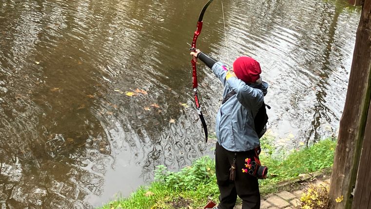 Une personne tire à l'arc sur la rive d'un plan d'eau. En arrière-plan, on peut voir des arbres et une surface d'eau calme.