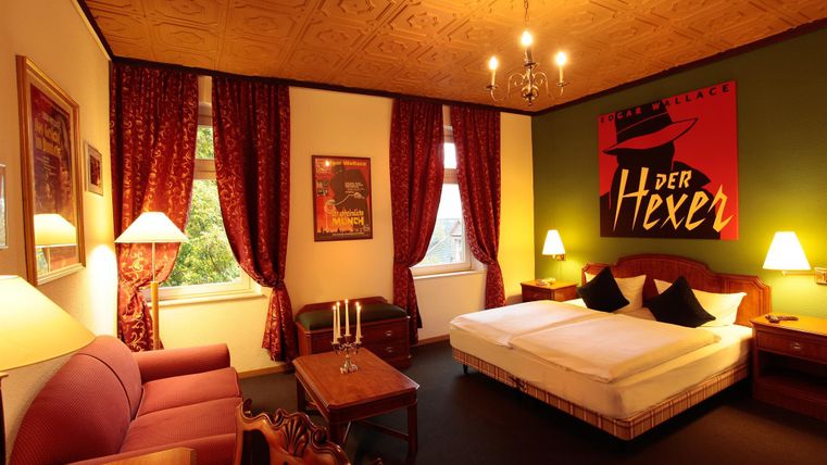 Une chambre d'hôtel confortable avec des rideaux rouges et un grand lit. Un œuvre d'art saisissante intitulée "le Sorcier" est accrochée au mur.